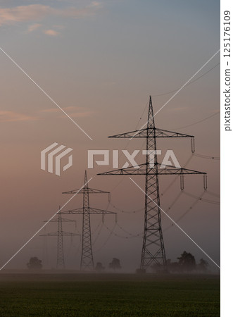 Electricity pylons in a row through fog at sunrise, in a german village Electricity pylons in a row through fog at sunrise, in a german village 125176109
