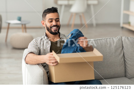 Quick delivery service concept. Happy young Arab guy receiving package, unpacking cardboard box, sitting on sofa in living room. Middle Eastern male buyer satisfied with online shopping purchase Quick delivery service concept. Happy young Arab guy receiving package, unpacking cardboard box, sitting on sofa in living room. Middle Eastern male buyer satisfied with online shopping purchase 125176123