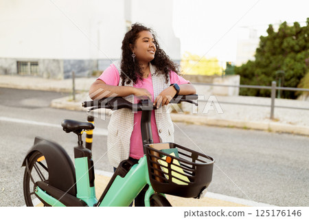 A woman leans on her electric scooter, gazing thoughtfully into the distance. She wears a pink shirt and a knitted vest, enjoying the warm afternoon sun on an urban street. A woman leans on her electric scooter, gazing thoughtfully into the distance. She wears a pink shirt and a knitted vest, enjoying the warm afternoon sun on an urban street. 125176146