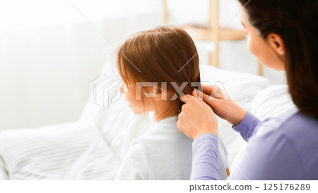 Mother care concept. Close up of woman braiding her little daughter hair, home interior 125176289