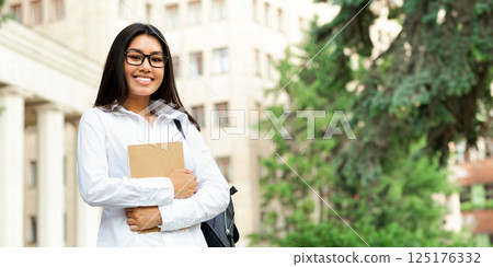 Asian college student holding books and smiling to camera in front of university Asian college student holding books and smiling to camera in front of university 125176332