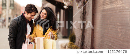 A couple enjoys their shopping experience, smiling and looking at their bags while walking down a bustling city street adorned with seasonal decorations, copy space 125176476