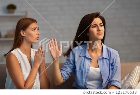 Miserable Girl Asking Indifferent Friend For A Favor Begging Holding Hands In Prayer Gesture Sitting On Sofa Indoor. Selective Focus 125176538