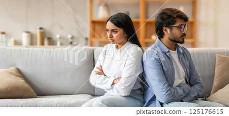 Indian couple sits on opposite ends of a couch, arms crossed and expressions serious, indicating a conflict. The warm interior reflects a sense of domestic life despite the tension. 125176615