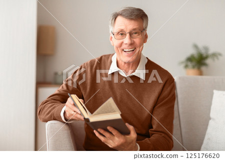 Positive Senior Man Holding Book Smiling At Camera Sitting On Sofa Indoor. Reading And Self-Development. Selective Focus 125176620