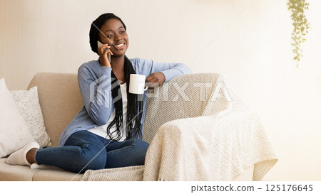 Smiling African American Woman Talking on Cellphone And Drinking Morning Coffee While Sitting On Couch At Home 125176645