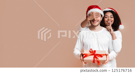 Black woman covering her boyfriend's eyes, giving him Christmas present and showing silence sign over yellow studio background with free space Black woman covering her boyfriend's eyes, giving him Christmas present and showing silence sign over yellow studio background with free space 125176716