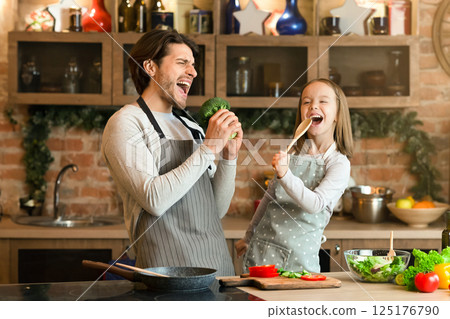 Father and daughter having fun while preparing lunch in kitchen, singing loud, using spatula and broccoli as microphones 125176790