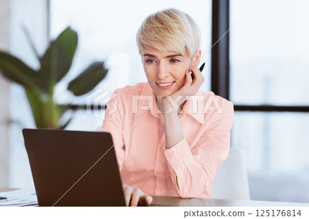 Smiling Mature Business Lady At Laptop Computer Working On A Project Sitting At Workplace In Office. Selective Focus Smiling Mature Business Lady At Laptop Computer Working On A Project Sitting At Workplace In Office. Selective Focus 125176814