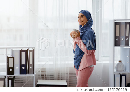 Break in work. Arabic female manager in hijab standing with coffee mug in office, relaxing against window at workplace, free space 125176919