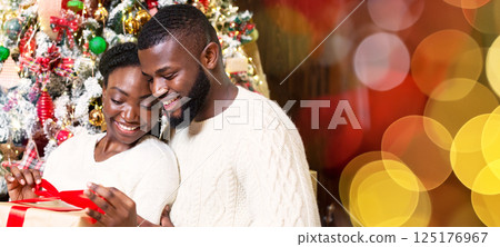 Happy african american woman opening Christmas gift box from her caring husband sitting near Xmas tree at home 125176967