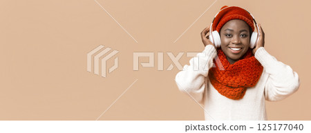 A cheerful young African American woman smiles while wearing a warm red scarf and stylish headphones. She is enjoying music in a cozy indoor environment, radiating happiness and comfort. 125177040