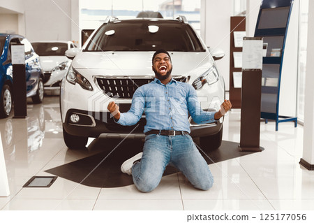 New Auto. Excited African Man Shouting Shaking Fists Celebrating Buying Car Standing On Knees In Dealership Store New Auto. Excited African Man Shouting Shaking Fists Celebrating Buying Car Standing On Knees In Dealership Store 125177056