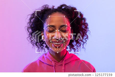 A young black woman with curly hair smiles while wearing wireless earbuds and experiencing facial recognition technology against a vibrant background. A young black woman with curly hair smiles while wearing wireless earbuds and experiencing facial recognition technology against a vibrant background. 125177159