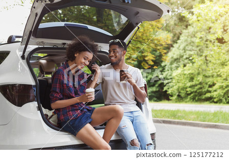 Happy afrocouple sitting in open car back, having stop off, drinking coffee. Romantic traveling concept Happy afrocouple sitting in open car back, having stop off, drinking coffee. Romantic traveling concept 125177212