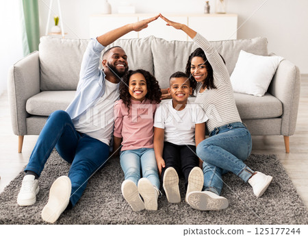 Family Care, Protection And Insurance Concept. Portrait of smiling African American parents making symbolic roof of hands above their happy children, sitting on the floor carpet in living room at home 125177244