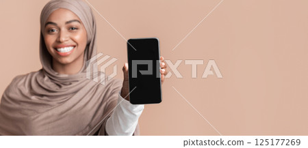 Modern smartphone with blank black screen in hand of smiling afro muslim girl in hijab, yellow background with empty space 125177269