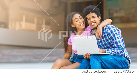 A teen black couple sits together on a ledge in the city, laughing and using a laptop. The girl is wearing a pink shirt and denim skirt, while guy wears a plaid shirt and jeans. A teen black couple sits together on a ledge in the city, laughing and using a laptop. The girl is wearing a pink shirt and denim skirt, while guy wears a plaid shirt and jeans. 125177330