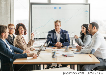 A group of business professionals are gathered around a table in a modern office, discussing a growth chart displayed on a large screen. The team is engaged in a lively conversation A group of business professionals are gathered around a table in a modern office, discussing a growth chart displayed on a large screen. The team is engaged in a lively conversation 125177332