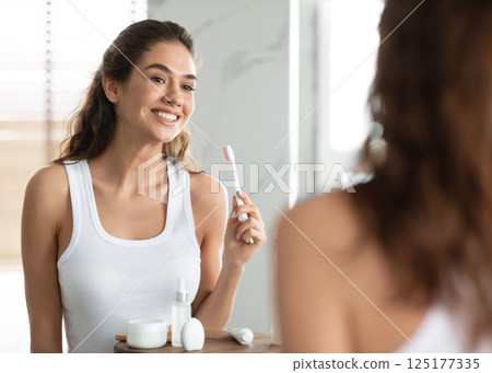 Toothcare. Happy Woman Brushing Teeth Standing Holding Toothbrush And Smiling To Mirror Reflection In Modern Bathroom At Home. Oral Hygiene Morning Routine, Clean Teeth And Perfect White Smile Concept 125177335
