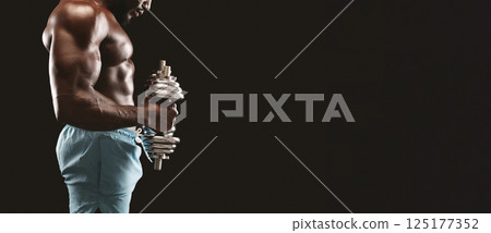 A muscular man in blue shorts lifts a heavy dumbbell while standing in a dark room. A muscular man in blue shorts lifts a heavy dumbbell while standing in a dark room. 125177352