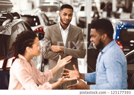 Car Seller Looking At Afro Couple Having Quarrel About Buying Too Expensive Car Standing In Dealership Showroom. Selective Focus 125177393