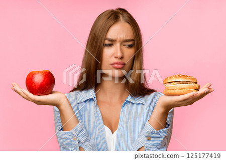 Puzzled Lady Holding Burger And Apple Choosing Between Unhealthy And Healthy Food Standing Over Yellow Background. Studio Shot Puzzled Lady Holding Burger And Apple Choosing Between Unhealthy And Healthy Food Standing Over Yellow Background. Studio Shot 125177419
