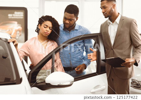 Displeased Car Buyers Looking At Auto Choosing New Automobile With Seller Man In Dealership Store. Selective Focus Displeased Car Buyers Looking At Auto Choosing New Automobile With Seller Man In Dealership Store. Selective Focus 125177422
