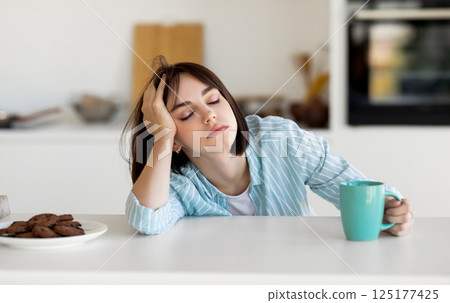 Sleepy young woman drinking coffee, feeling tired, suffering from insomnia and sleeping disorder. Sad female sitting in modern kitchen interior, empty space Sleepy young woman drinking coffee, feeling tired, suffering from insomnia and sleeping disorder. Sad female sitting in modern kitchen interior, empty space 125177425