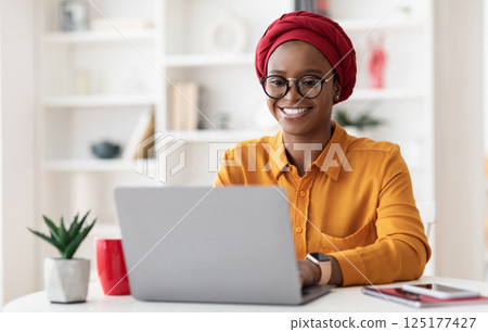 Cheerful millennial black woman entrepreneur working from home, wearing red headscarf and eyeglasses, sitting at workdesk and typing on laptop keyboard, chatting with clients, copy space 125177427