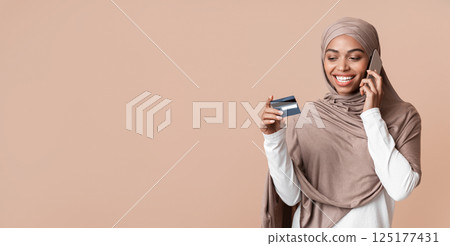 Customer service concept. Young black muslim woman in hijab holding credit card and talking on cellphone with bank manager, yellow background Customer service concept. Young black muslim woman in hijab holding credit card and talking on cellphone with bank manager, yellow background 125177431