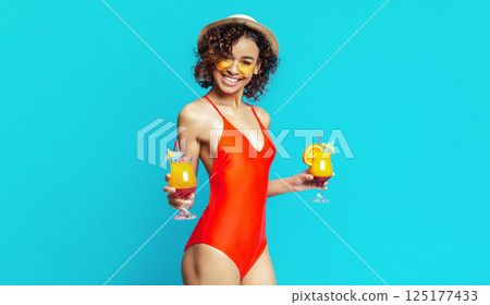 Black woman in a red swimsuit, holding two cocktails with orange slices and umbrellas. She is smiling and standing against a bright blue background, wearing a white hat and yellow sunglasses. 125177433