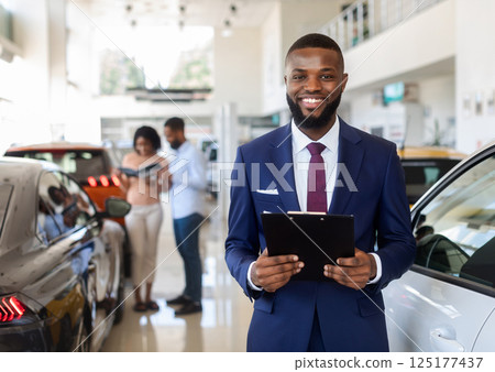 Portrait Of Handsome Black Car Salesman In Suit Posing At Workplace In Auto Showroom, Young Dealership Center Manager With Clipboard In Hands Helping Spouses To Purchase New Automobile, Free Space Portrait Of Handsome Black Car Salesman In Suit Posing At Workplace In Auto Showroom, Young Dealership Center Manager With Clipboard In Hands Helping Spouses To Purchase New Automobile, Free Space 125177437