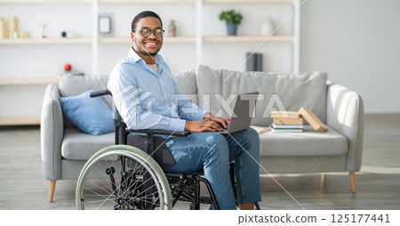 Happy disabled black man in wheelchair using laptop, working online from home, copy space. Millennial African American handicapped guy having business or studying on web 125177441