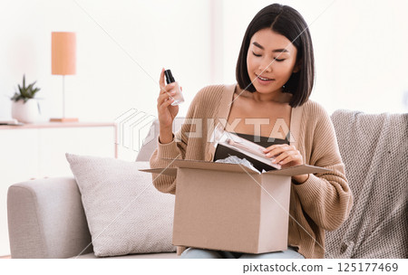 Online Shopping. Smiling asian woman opening parcel box with cosmetics at home, holding makeup products, unpacking gift sitting on the couch at home. Lady satisfied with delivery service 125177469
