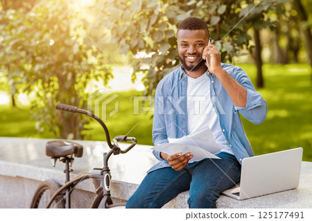 Out Of Office. Smiling Black Businessman With Papers And Laptop Talking On Cellphone With Clients, Sitting On Bench In Park Out Of Office. Smiling Black Businessman With Papers And Laptop Talking On Cellphone With Clients, Sitting On Bench In Park 125177491