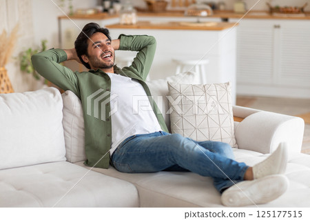 Arab man in a green button-down shirt and white t-shirt reclines on a white couch, his arms behind his head, smiling and looking up. 125177515