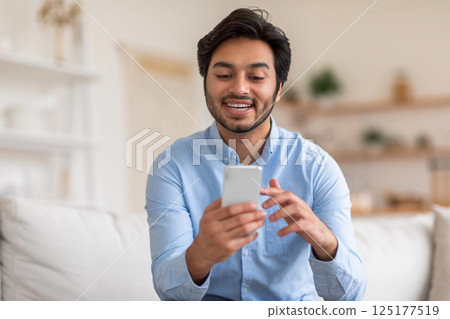 A young Arab man with short dark hair and a blue button-up shirt is sitting on a light colored couch. He is using his smartphone, looking at the screen, and smiling. A young Arab man with short dark hair and a blue button-up shirt is sitting on a light colored couch. He is using his smartphone, looking at the screen, and smiling. 125177519