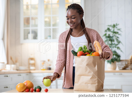 Black Smiling Lady Unpacking Paper Bag With Fresh Organic Vegetables And Fruits After Grocery Shopping, Happy African American Woman Standing In Cozy Kitchen Interior At Home, Copy Space Black Smiling Lady Unpacking Paper Bag With Fresh Organic Vegetables And Fruits After Grocery Shopping, Happy African American Woman Standing In Cozy Kitchen Interior At Home, Copy Space 125177594