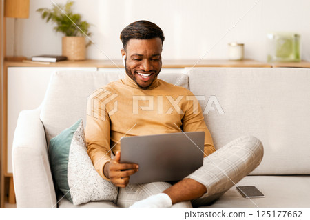 Joyful African American Man Using Laptop And Earbuds Making Video Call Sitting On Couch At Home. Free Space, Selective Focus 125177662