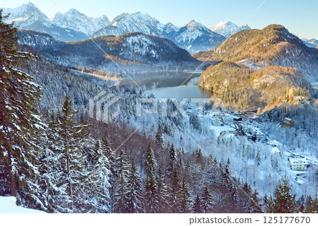 Snowy Mountain Landscape with Iconic Castle Overlooking Serene Lake and Alpine Peaks in Ethereal Winter Wonderland Bathed in Soft Dawn Light 125177670