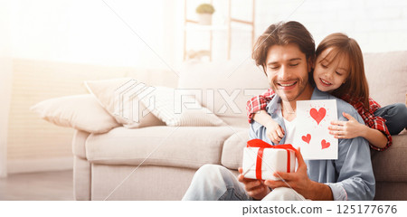 Cute preschool girl greeting her happy daddy with Father Day, giving him gift box and greeting card, home interior, free space 125177676