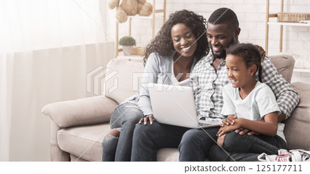 African american family of three using laptop together while sitting on sofa at home, watching videos. 125177711