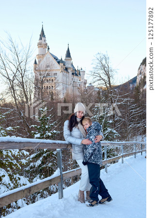 Beautiful woman and child standing on a bridge in the snow with Neuschwanstein Castle in the background, capturing a serene winter scene with a majestic castle and snowy landscape. 125177892