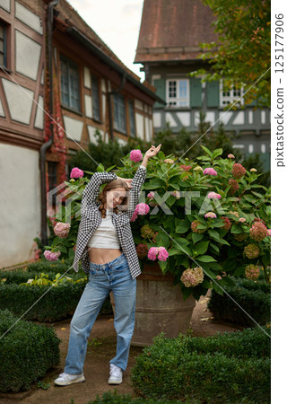 Stylish Young Woman in Checkered Shirt and Crop Top Poses with Pink Hydrangeas by Half-Timbered Houses. Carefree Fashion Portrait Captures Rural Village Charm and Vibrant Fall Foliage in Picturesque Stylish Young Woman in Checkered Shirt and Crop Top Poses with Pink Hydrangeas by Half-Timbered Houses. Carefree Fashion Portrait Captures Rural Village Charm and Vibrant Fall Foliage in Picturesque 125177906