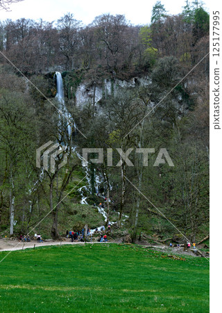 Serene Lush Green Meadow With Winding Dirt Path Leading To Cascading Waterfall Amidst Rocky Cliff And Leafless Trees Under Overcast Sky In Early Spring. Serene Lush Green Meadow With Winding Dirt Path Leading To Cascading Waterfall Amidst Rocky Cliff And Leafless Trees Under Overcast Sky In Early Spring. 125177995