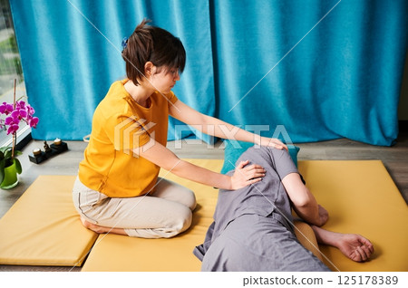 Masseuse making thai yoga massage. Woman therapist doing traditional massage treatment for male patient indoors. Bald client lying on mat, meditating, enjoying hand massage in calm atmosphere. 125178389