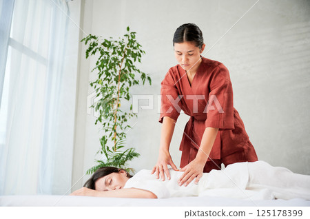 Masseuse making thai yoga massage. Woman therapist doing traditional massage treatment for female patient indoors. Client lying on mat, meditating, enjoying hand massage in calm atmosphere. 125178399