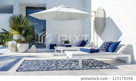 A tranquil outdoor lounge area features white lounge chairs and an umbrella, perfect for relaxation. Blue cushions and a patterned rug enhance the serene atmosphere near a swimming pool. 125178599