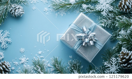 A silver gift box adorned with a ribbon sits at the center, surrounded by pine branches, pinecones, and artificial snowflakes. The serene blue background creates a winter wonderland atmosphere. 125179620
